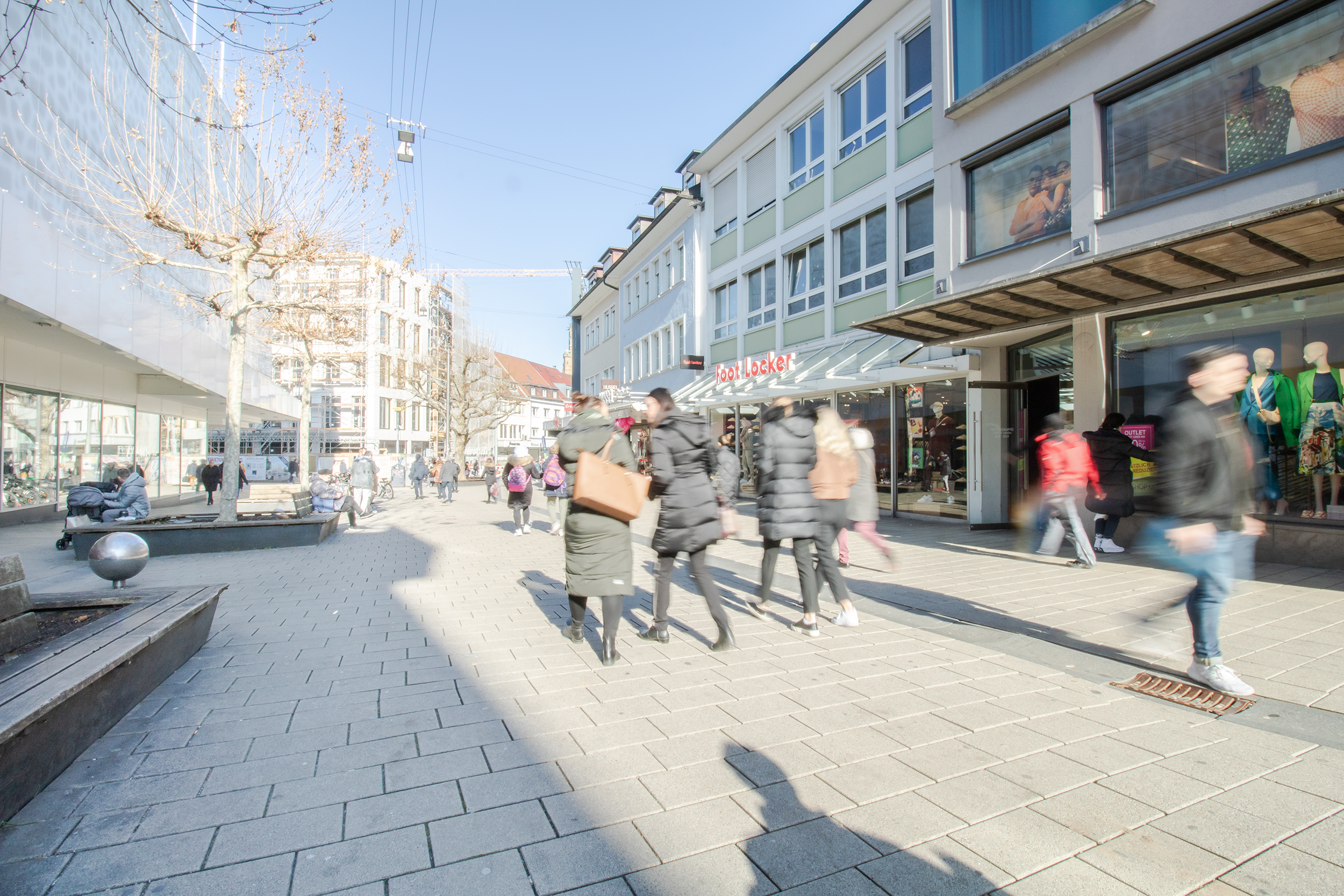 Das Bild zeigt die Fußgängerzone mit dem Blick Richtung Innenstadt. Links im Bild ist das Einkaufszentrum Galeria Kaufhof zu sehne. Rechts sind weitere kleine Läden abgebildet. Passanten laufen die Fußgängerzone entlang.